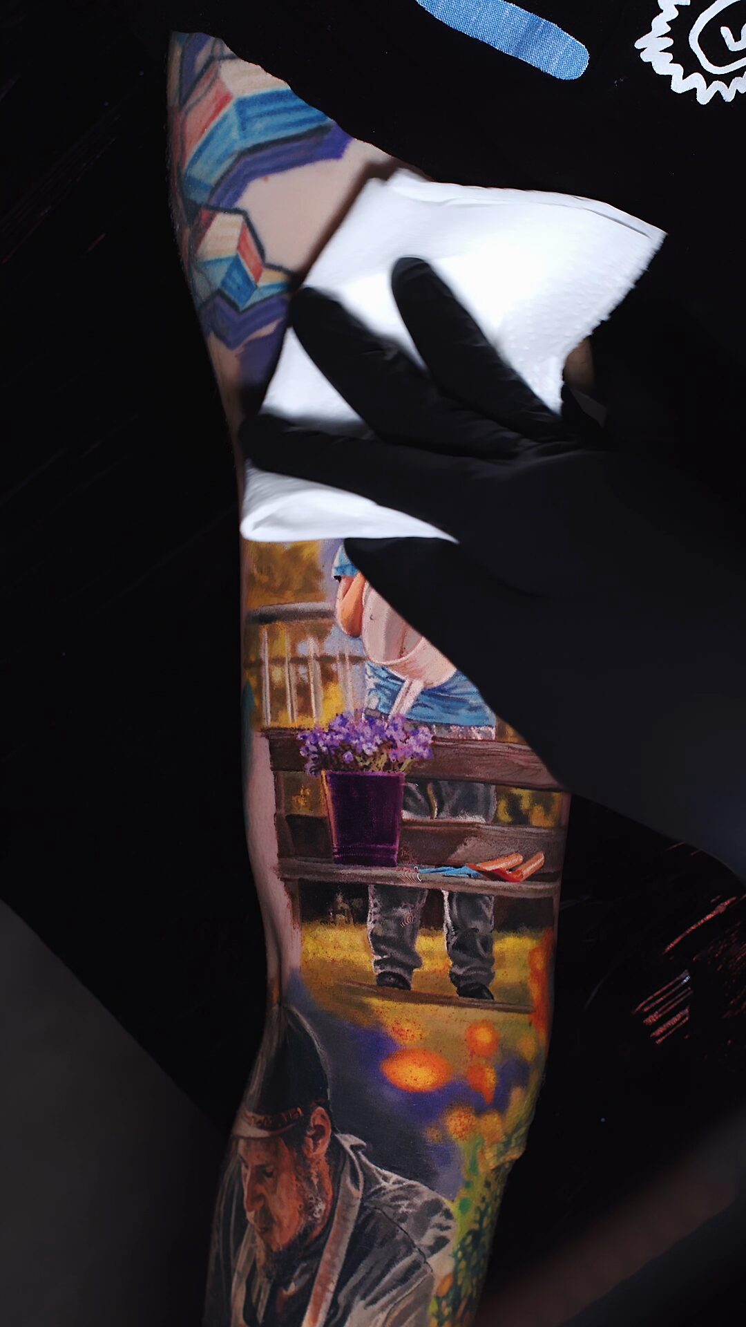 Das komplette Tattoo am Bizeps: Junge mit Gießkanne, lila Blumen, Holzzaun, Berge im Hintergrund – Color-Realism