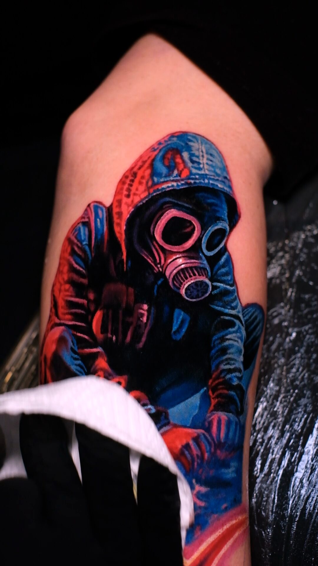 Tattoo einer Figur mit Kapuze und Gasmaske auf dem Oberarm – Seitenansicht, Handtuch unten, Wrocław.
